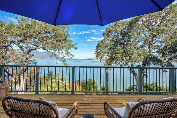 A wooden deck with wicker chairs and a blue umbrella, overlooking a lake with trees and mountains in the background. - Views! Private! Tranquil! Lakefront! Forest! Sunsets! Acreage! Docks! (Lower Lake)