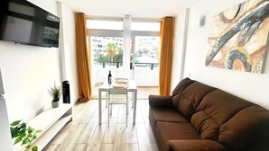 Ein modernes Wohnzimmer mit einem braunen Sofa, einem an der Wand montierten Flachbildfernseher, einem Esstisch mit Stühlen und einem großen Fenster mit Vorhängen.