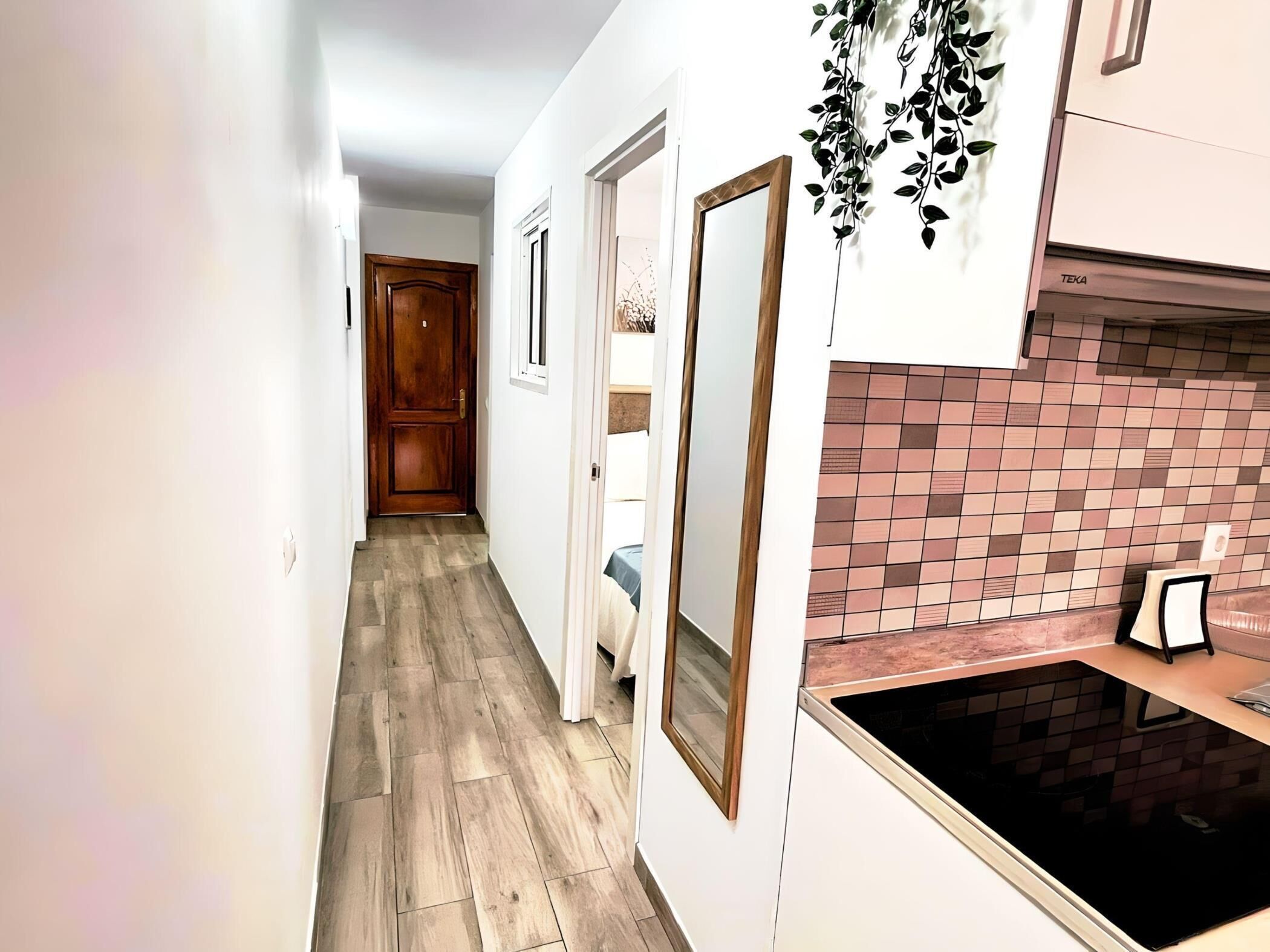 A narrow hallway with wooden flooring, white walls, and a kitchen area with a black countertop and tiled backsplash.