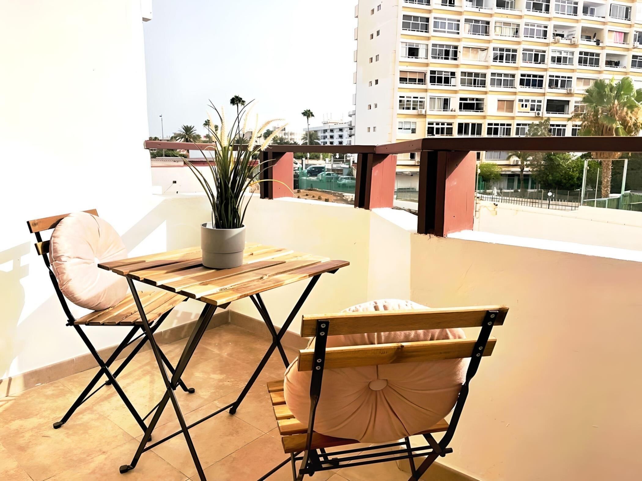 A balcony with a wooden table and two chairs, a potted plant, and a view of a residential building.