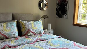 Un dormitorio con una colcha colorida, un espejo redondo, una lámpara de noche y una ventana con vistas a árboles.