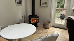 Una habitación acogedora con una mesa blanca, sillas de mimbre, un estufa de leña negra y cuadros enmarcados en la pared.