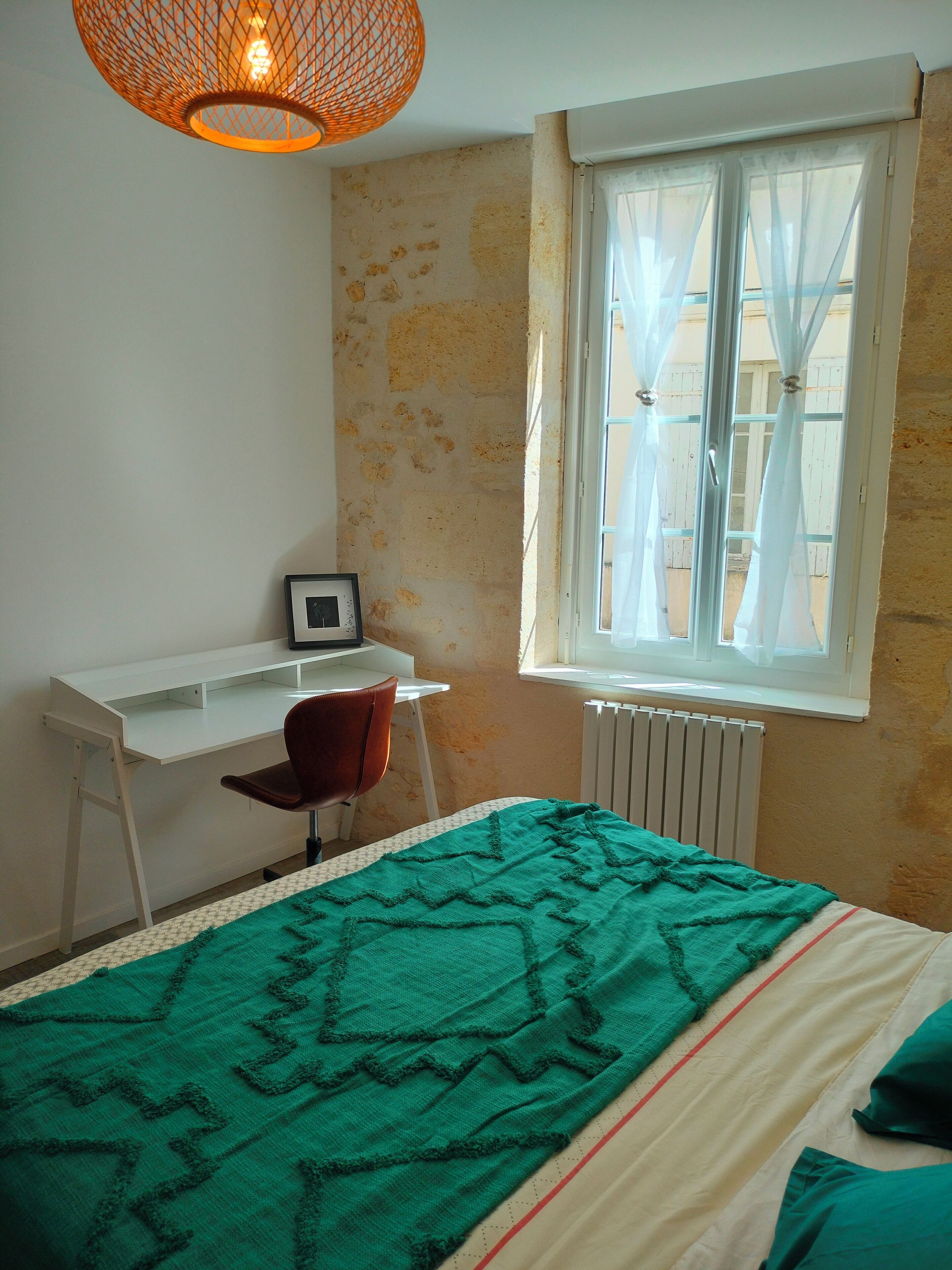 Een slaapkamer met een bed, een bureau, een stoel, een raam met gordijnen en een radiator.