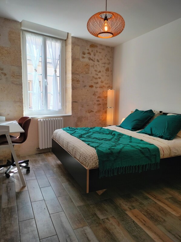Un dormitorio con cama, escritorio, silla, ventana con cortinas y lámpara colgante.
