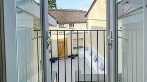 Udsigt gennem en glasdør, der fører til en balkon med gelænder, en trædør og en bygning med stråtækt tag i baggrunden.