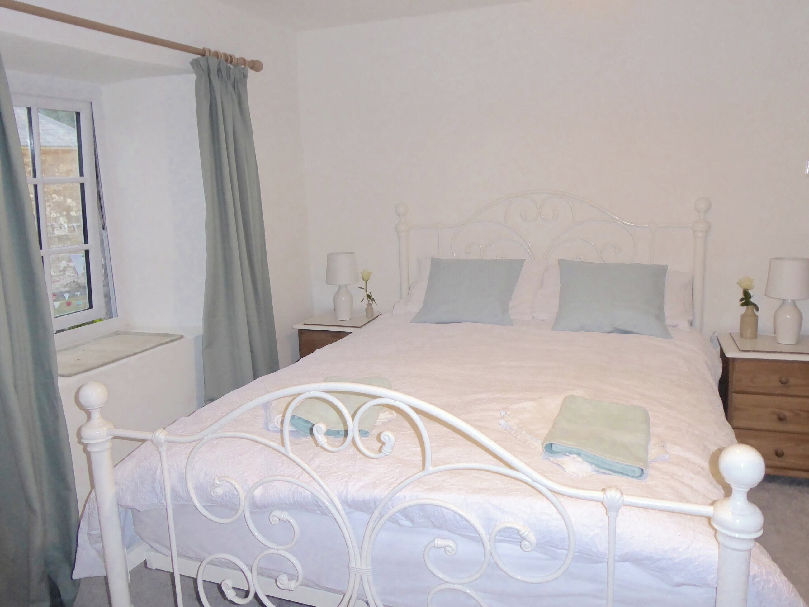 A neatly made bed with a white metal frame, a bedside table with a lamp, and a window with curtains.