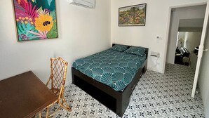 Un dormitorio con una cama, una silla, una mesa, un aire acondicionado y dos cuadros en la pared.