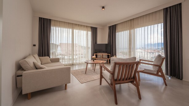 A modern living room with a sofa, two wooden chairs, a small coffee table, and a TV. Large windows with curtains provide natural light and a view of the city. - Gizz Suites City (Fethiye)