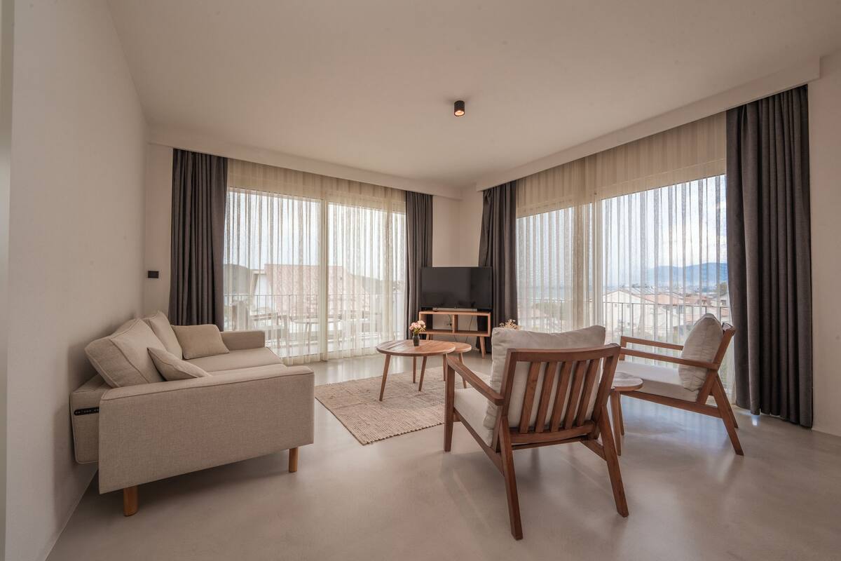 A modern living room with a sofa, two wooden chairs, a small coffee table, and a TV. Large windows with curtains provide natural light and a view of the city.