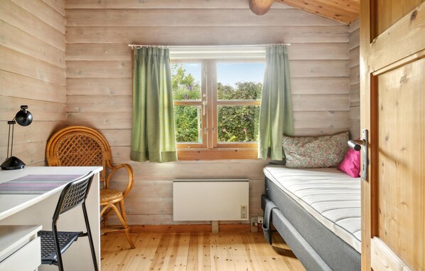 A small, cozy room with wooden walls, a window with green curtains, a bed with a floral pillow, a chair, and a desk with a lamp. - Amazing home in Stege with kitchen (Stege)