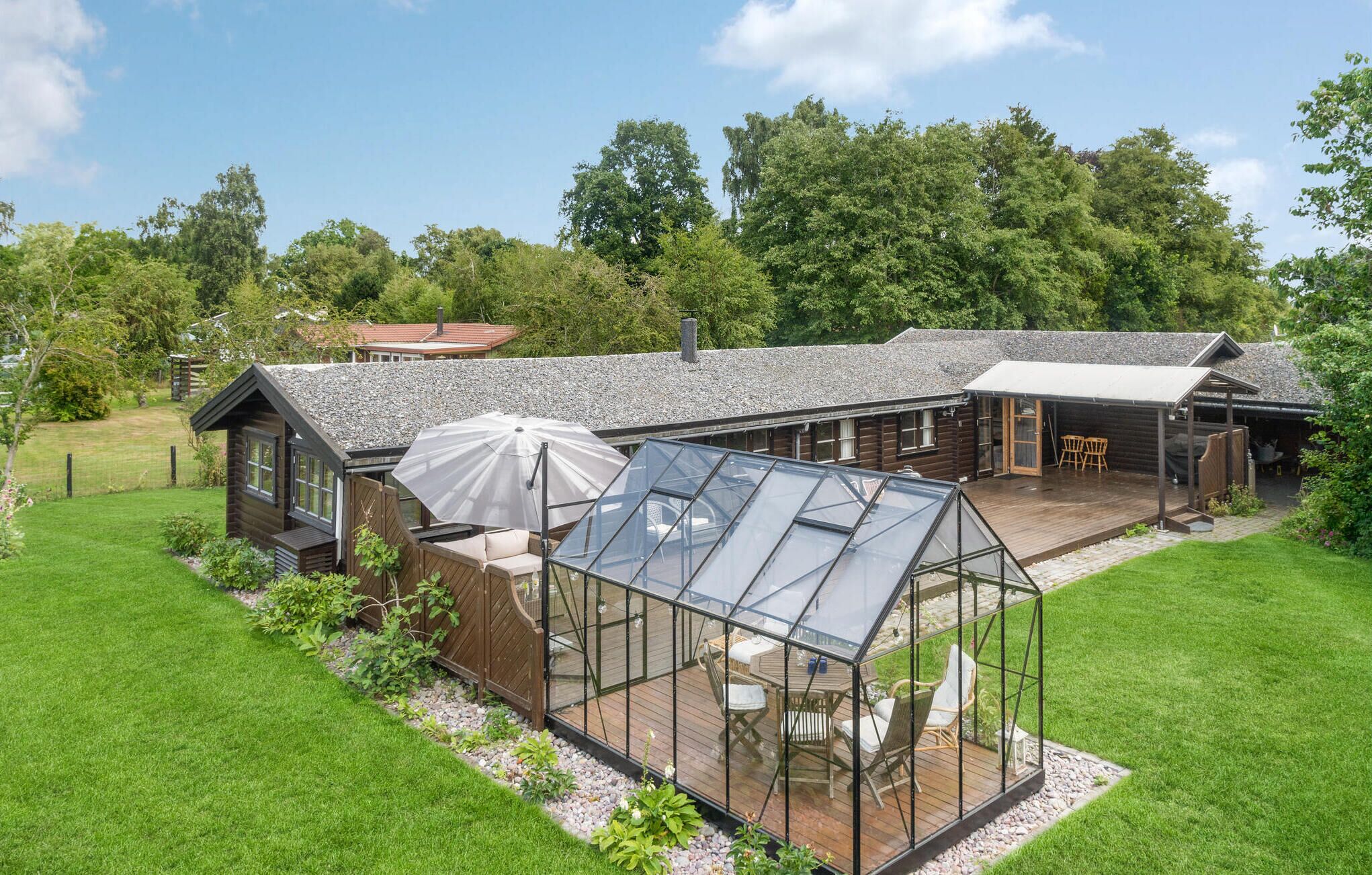 Une maison dotée d’une grande serre en verre, d’une terrasse en bois et d’une cour clôturée.