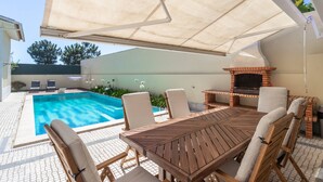 Un espace aménagé au bord de la piscine, avec une table et des chaises en bois, un barbecue et un coin salon abrité.
