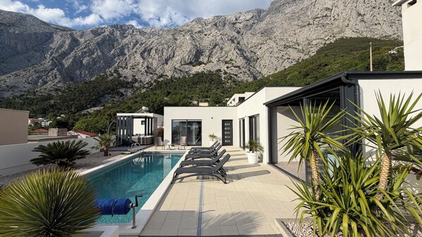 A modern pool area with a mountain backdrop, including a pool, lounge chairs, and a small structure with a glass door.