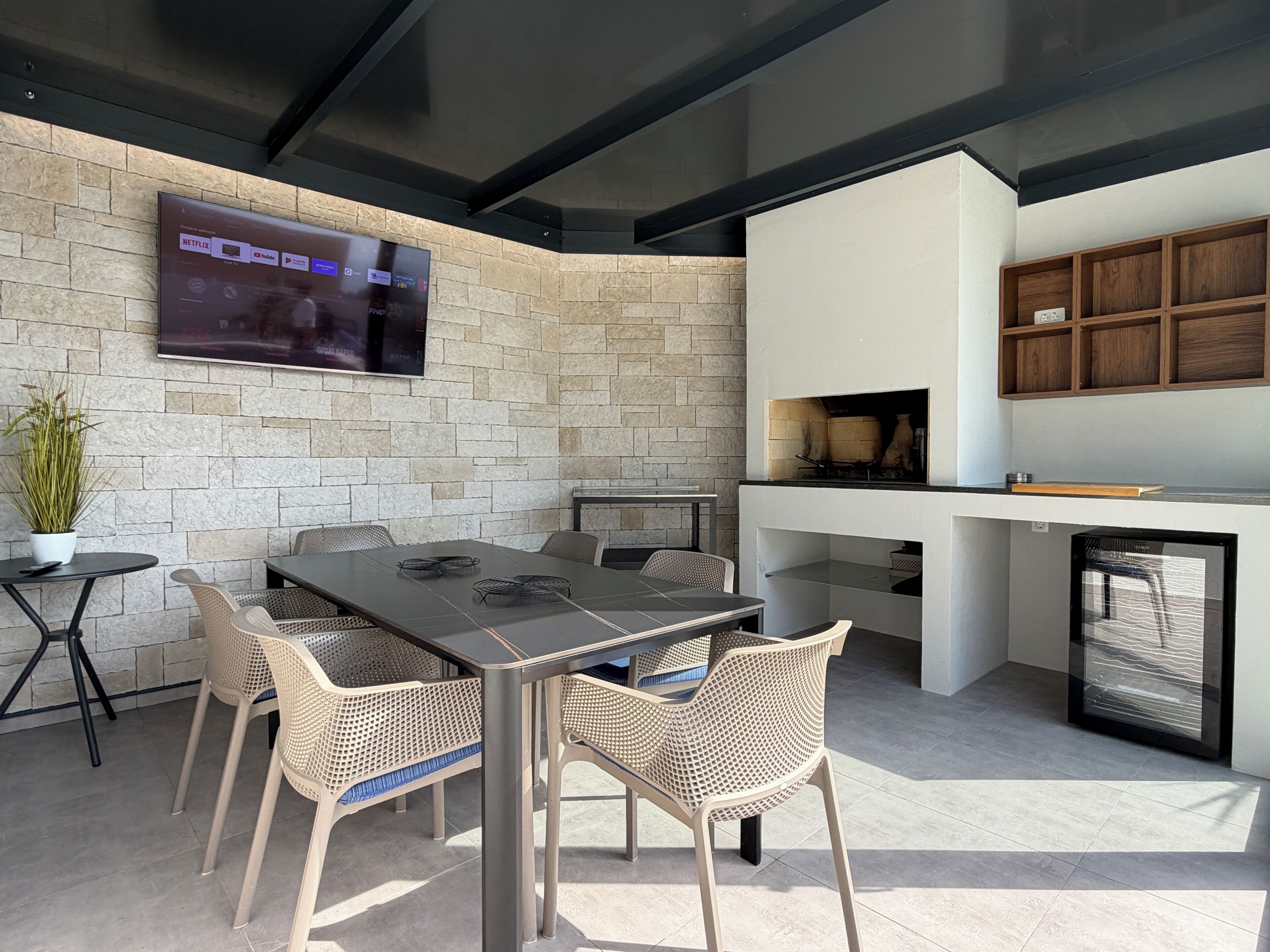 Un espace de restauration extérieur moderne, agrémenté d’un mur en pierre, d’un téléviseur à écran plat fixé au-dessus d’une table, de chaises en osier et d’un barbecue encastré.