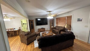 A living room with a wooden floor, a brown sofa, a coffee table, a TV, and a ceiling fan. - Ben Ranch House overlooking river - Full 3 Bedroom house. Minutes from Fairfax. (Ralston)
