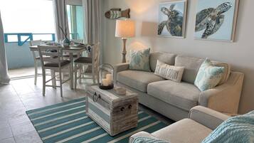 A living room with a sofa, a coffee table, and a striped rug. There is a dining table with chairs and a window with curtains.