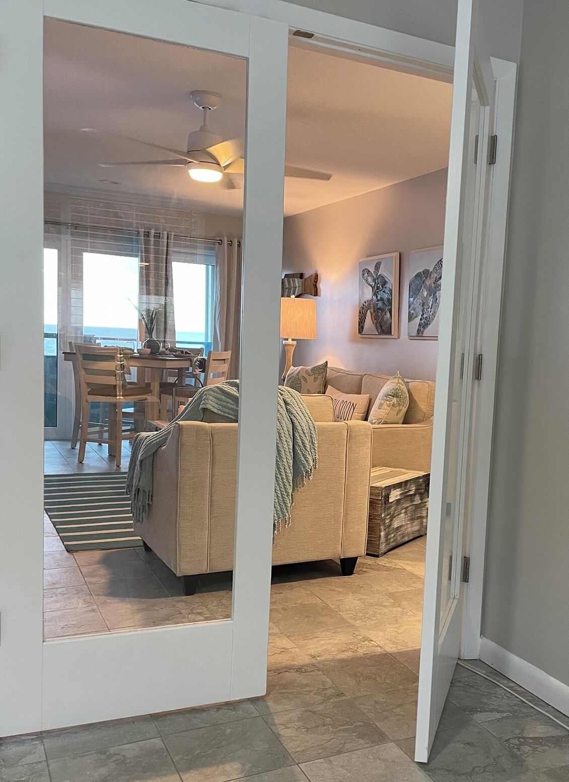 A modern living room with a beige sofa, a dining area with a table and chairs, and a view of the ocean through large windows.
