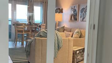 A modern living room with a beige sofa, a dining area with a table and chairs, and a view of the ocean through large windows.