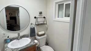 Ein Badezimmer mit einem weißen Waschbecken, einem Spiegel und einer Toilette. Darüber befindet sich ein Ablagefach und daneben ein Fenster.