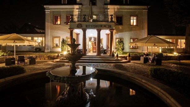 A large building with a sign reading "RESTAURACJA" at the entrance, surrounded by a well-lit outdoor area with a fountain and seating. - Pałac na Wodzie  (Podgorzyn)