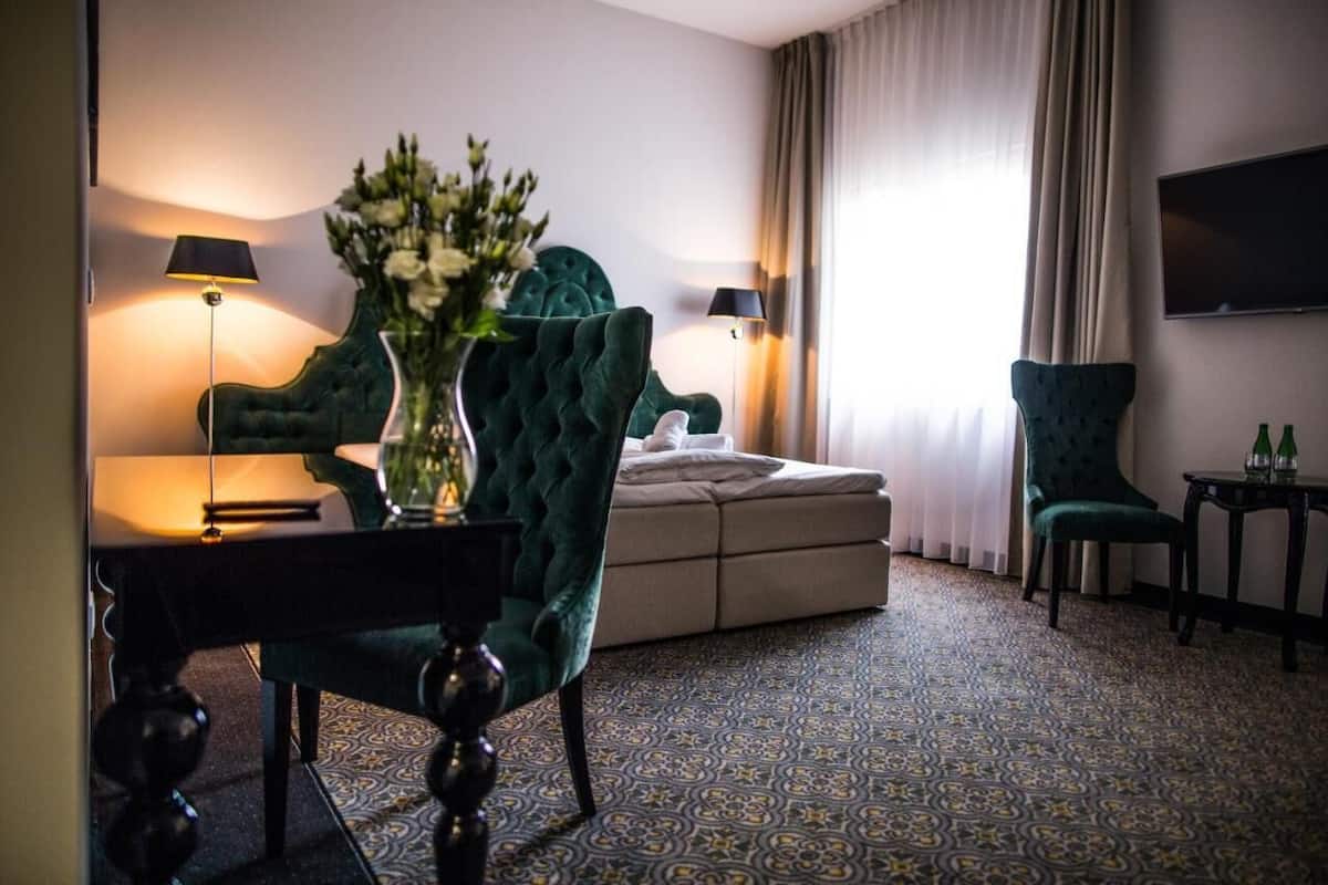 A hotel room with a green armchair, a sofa, a small table, a vase with flowers, and a television mounted on the wall.