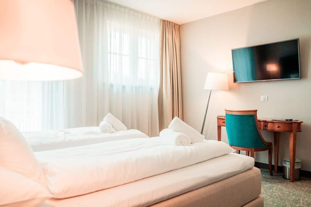 A hotel room with two beds, a desk, a chair, a TV, and a window with curtains.