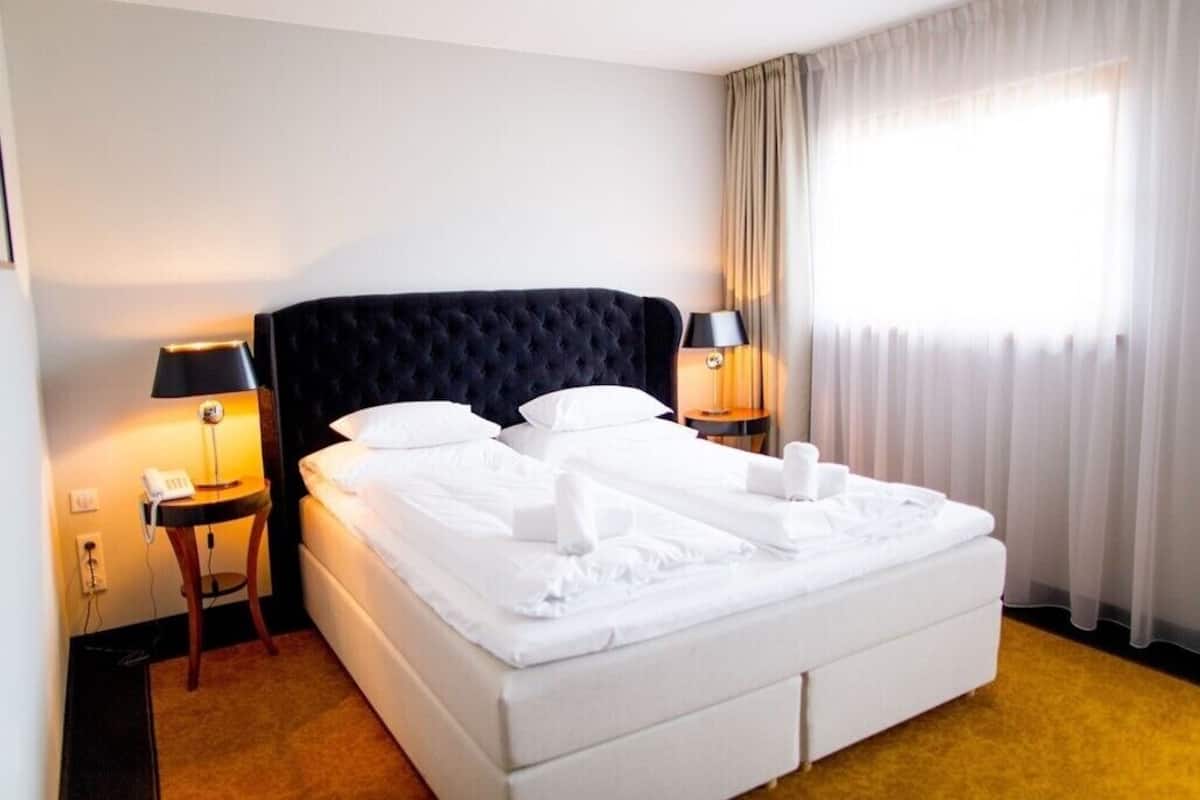 A hotel room with a large bed, two bedside tables with lamps, a telephone, and a window with curtains.