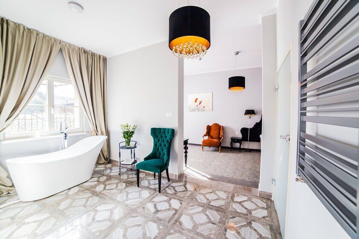 A modern bathroom with a freestanding bathtub, a green armchair, and a large window with beige curtains.