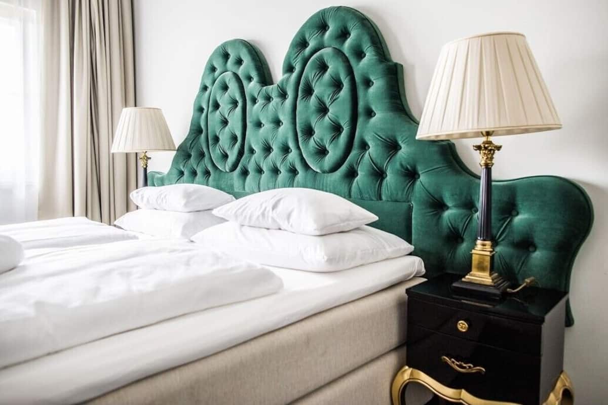 A neatly made bed with white linens and pillows, a green upholstered headboard, and a black nightstand with a gold lamp.