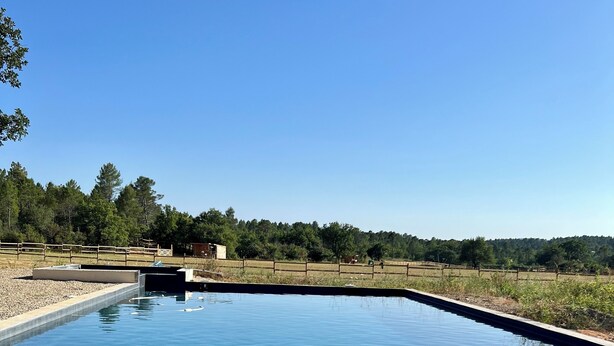A clear blue swimming pool with a wooden deck and a stone border. - le Charme D'une Cabane, le Confort en Plus (Aups)