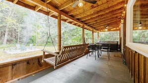 A wooden deck with a swing, outdoor seating, and a grill area, surrounded by trees.