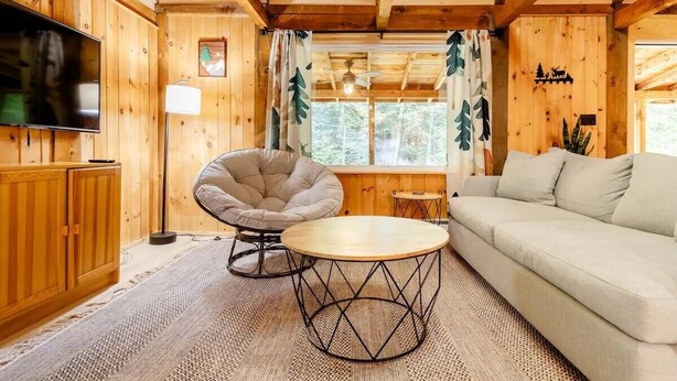 A cozy wooden cabin interior with a sofa, a round table, a chair, and a television.