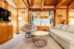 A cozy wooden cabin interior with a sofa, a round table, a chair, and a television.