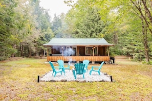 A cabin with a covered porch, surrounded by trees. There are four turquoise Adirondack chairs in front of the cabin.