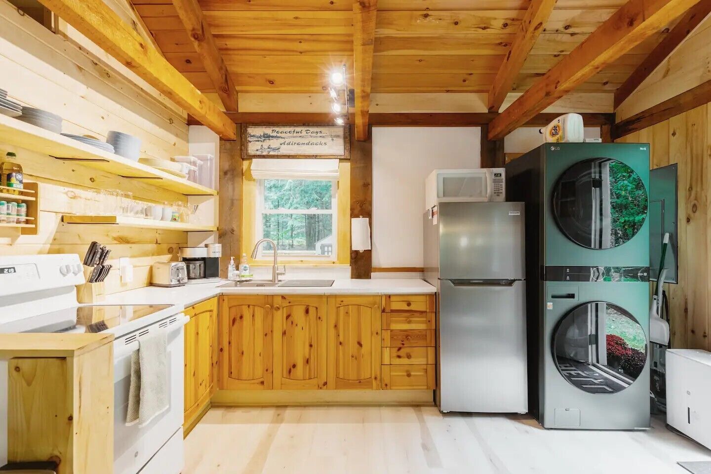 A compact kitchen with wooden cabinets, a stainless steel refrigerator, a washing machine, and a window providing a view of the outdoors.