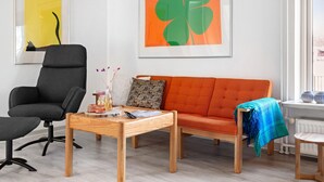 A modern living room with a grey office chair, an orange sofa, and a wooden coffee table. There are two framed artworks on the wall, a window with a view, and a radiator. - 2 bedroom amazing home in Sørvad (Sørvad)