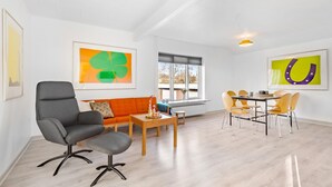 A modern living room with a grey armchair, orange sofa, wooden coffee table, and a dining area with yellow chairs. - 2 bedroom amazing home in Sørvad (Sørvad)