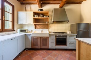 Una cocina compacta con armarios blancos, estufa a gas, horno empotrado, refrigerador y ventana con persianas.