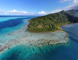 An aerial view of a tropical island with clear turquoise waters, dense green vegetation, and a small settlement with visible structures. - LTB FENUA Farm & Camping (Raiatea)