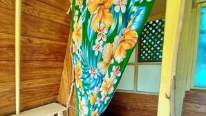 Une pièce avec un mur en bois, un banc, un lit agrémenté d’un rideau à motifs floraux verts et une petite fenêtre ornée d’un croisillon.
