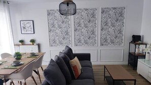 Ein modernes Wohnzimmer mit einem grauen Sofa, einem Holzespeisetisch und einem schwarzen Couchtisch. Hinter dem Sofa befinden sich zwei große, schwarz-weiß gemusterte Wandpaneele.