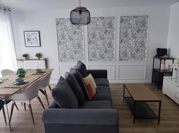 Ein modernes Wohnzimmer mit einem grauen Sofa, einem Holzespeisetisch und einem schwarzen Couchtisch. Hinter dem Sofa befinden sich zwei große, schwarz-weiß gemusterte Wandpaneele.
