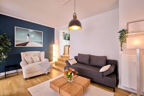Uma sala de estar com um sofá bege, um sofá cinza escuro, uma mesa de centro de madeira com um vaso de flores, uma fotografia emoldurada de um farol e um letreiro com a inscrição "EAT SLEEP TRAVEL REPEAT".