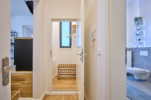 Un pasillo con un banco de madera, una puerta con manija y vista a un cuarto con inodoro y radiador.
