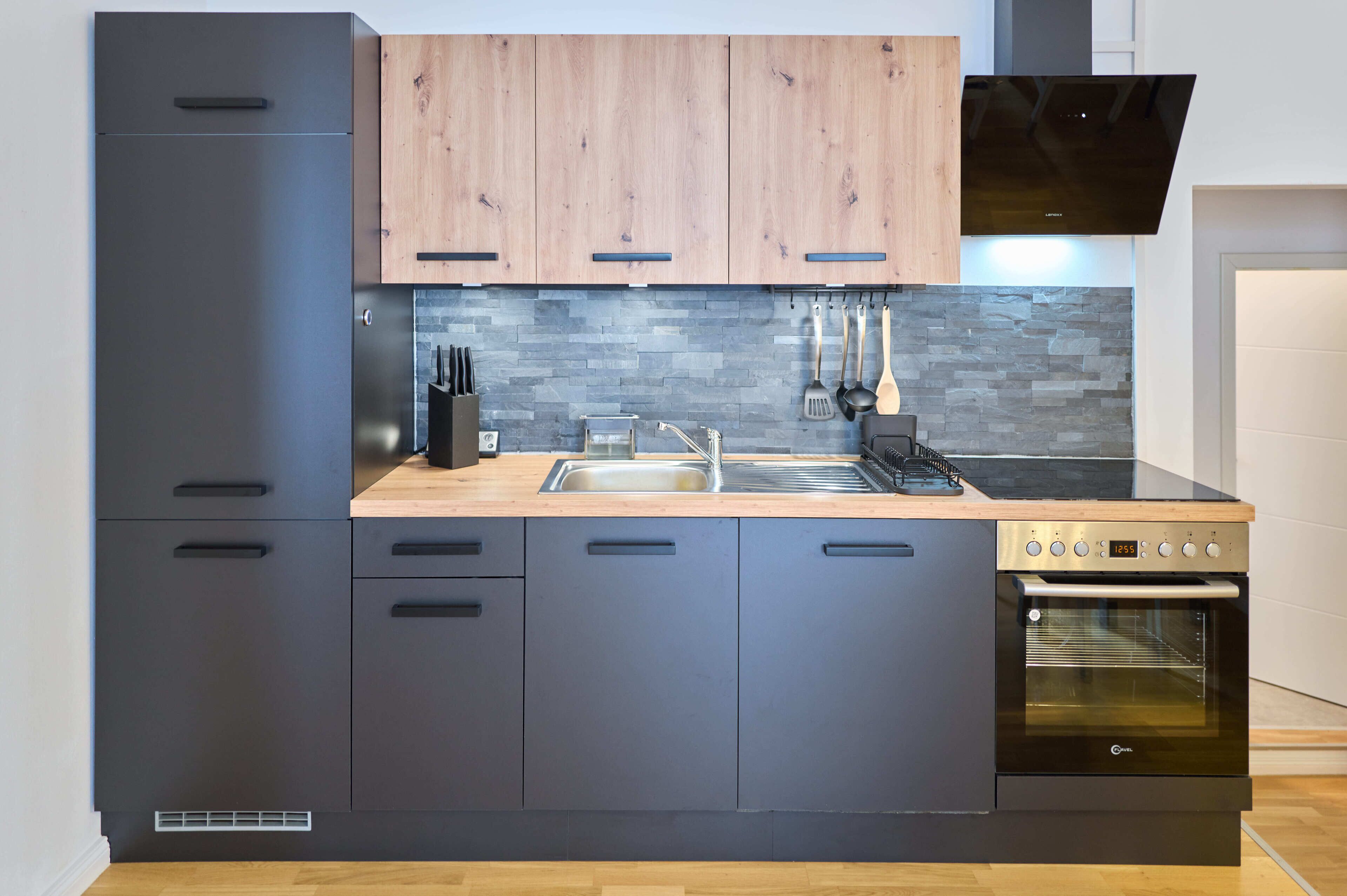 Une cuisine moderne avec des meubles de couleur sombre, un plan de travail en bois, un four encastré et un pare-feu en pierre.