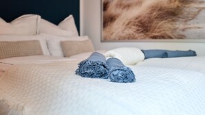 Een net opgemaakt bed met opgerolde handdoeken, een wit geweven sprei en een groot abstract schilderij aan de muur.