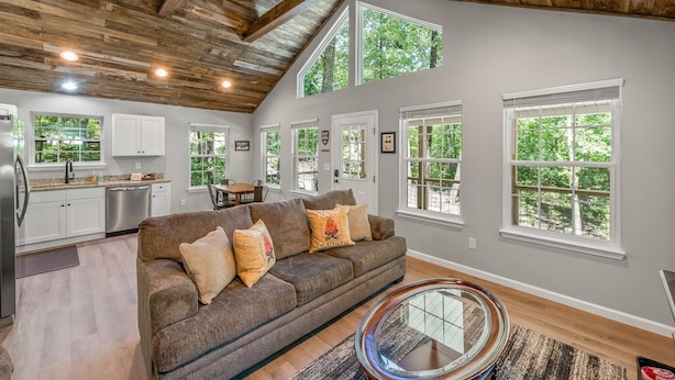 A spacious living area with a wooden ceiling, a brown sofa with decorative pillows, a glass coffee table, and large windows offering a view of greenery. - The Otter Box Cabin on 92 acres beside DeSoto State Park | Walking Ramp | Wi-Fi (Fort Payne)