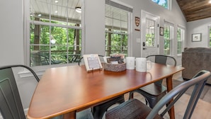 A dining area with a wooden table, chairs, and a view of greenery through large windows. - The Otter Box Cabin on 92 acres beside DeSoto State Park | Walking Ramp | Wi-Fi (Fort Payne)