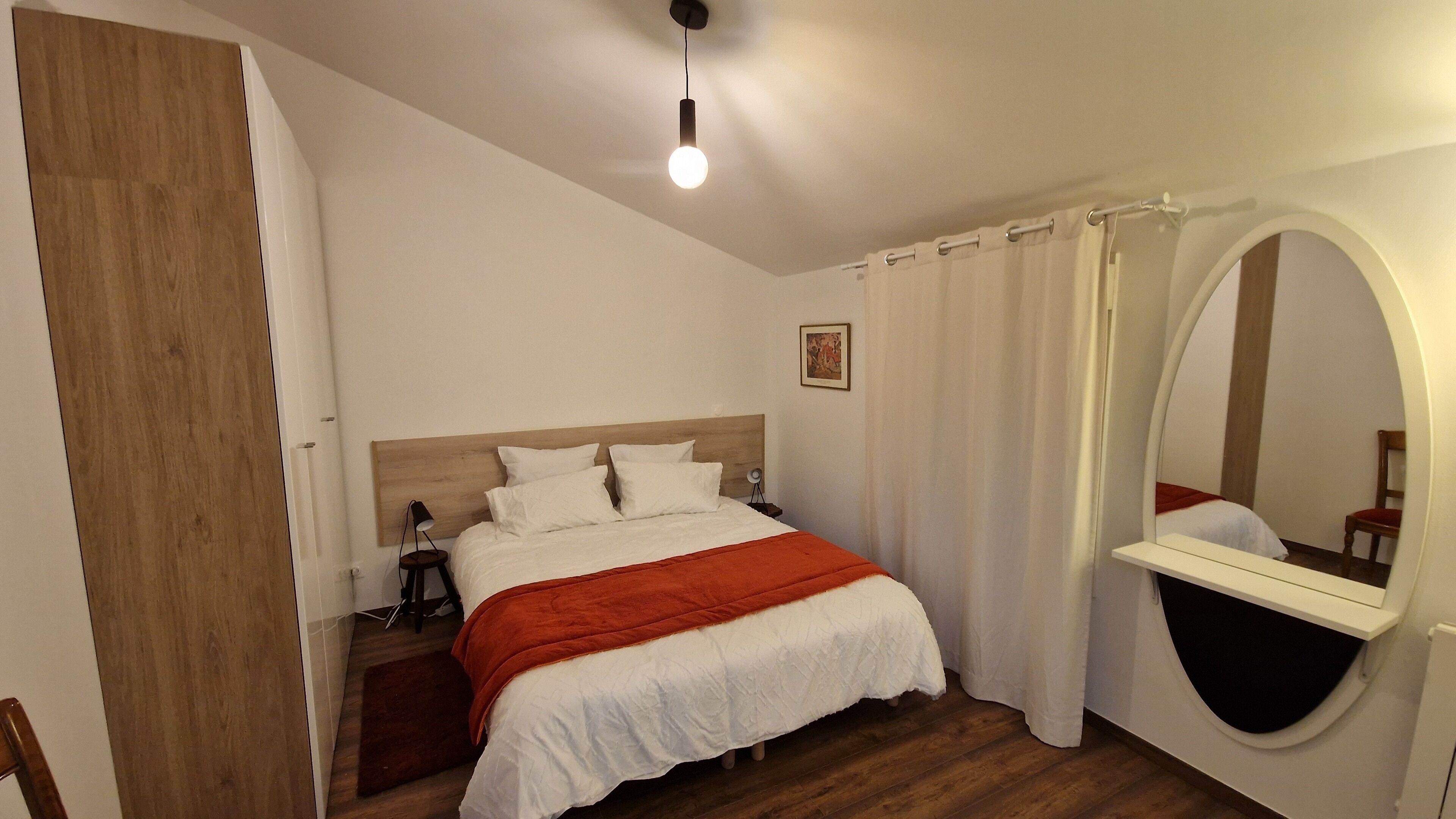 Un dormitorio con cabecera de madera, cama con ropa de cama blanca y una manta roja, mesita de noche con lámpara, un espejo grande y suelo de madera.