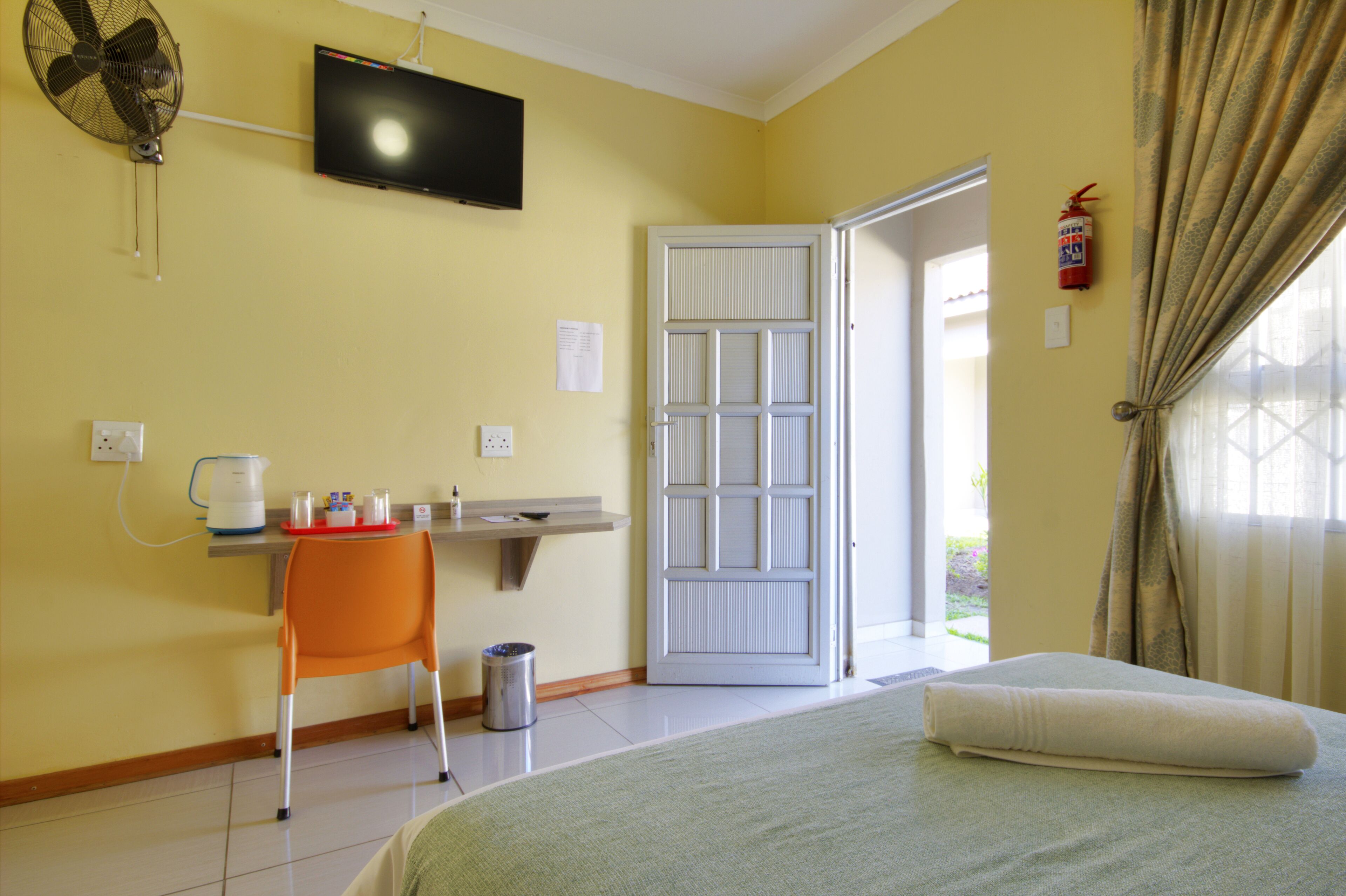 A hotel room with a bed, a desk with a chair, a television, a door, and a window with curtains.